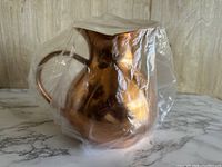 Front side of vintage copper pitcher wrapped in clear plastic showing pear-shaped round body with narrow mouth and handle