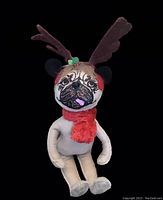 Front view of plush pug with reindeer antlers, red scarf, and earmuffs against black background.