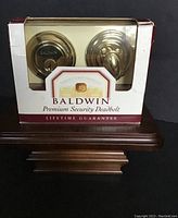 Front view of Baldwin Premium Security Deadbolt deadbolt in original packaging showing solid polished brass finish and visible lock components.