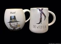 Front view of two mugs showing 'blessed' Pilgrim and 'The Witch is In' witch legs text and illustrations.