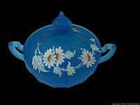 Side view of the blue opaque glass two-handle jar with painted daisies, lid with pointed finial, and visible crack on one handle.