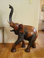 Full side profile of a large wooden carved elephant with raised trunk, showing tusks and detailed carved darker accents on trunk and legs.