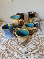 Group photo of six ceramic mugs and one ceramic pitcher arranged on lace cloth, showing varied glaze colors of blue, brown, cream, and earth tones.