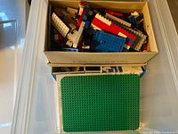 Box containing red, blue, white Lego bricks and pieces, plus large green baseplate beneath.