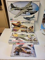 Four vintage airplane model kits stacked: B-17F Memphis Belle, Spitfire Mk.1a, Bf 109E, SPAD XIII biplane. Box art shows detailed paint schemes and scale.
