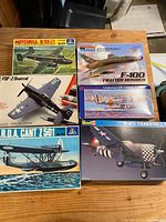 Lot of six vintage plastic model airplane kits on wooden table, showing illustrated box art for Mitchell B-25 bomber, F-100 fighter bomber, P-47 Thunderbolt, F8F-2 Bearcat, Grumman J2F-2 Duck, and Cant Z501 seaplane