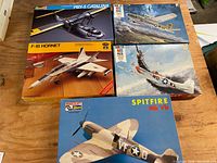 Four boxed vintage model airplane kits and two aircraft puzzles displayed on a wooden surface.