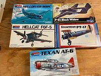 Five vintage plastic model airplane kits arranged on wooden surface including Monogram Helldiver SB2C, Hellcat F6F-5, Texan AT-6, MPC P-61 Black Widow, and Stearman PT-17 boxes.