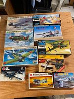 Lot of vintage model airplane kits displayed on wood surface showing box art and titles, highlighting Japanese WW2 planes and others.