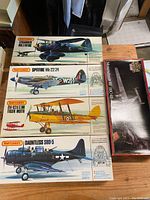 Four vintage Matchbox model airplane kits laid flat on a wooden table showing the top of each box: Lysander Mk I/III, Spitfire Mk 22/24, Tiger Moth, Dauntless SBD-5, plus a separate boxed model of a sword from The Lord of the Rings series.