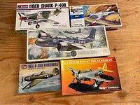 Five vintage plastic model airplane kits on a wooden surface including Monogram Tiger Shark P-40B, Hasegawa Shooting Star T-33A, Airfix Douglas Invader, Airfix Bell P-390 Airacobra, and Heller Humbrol Republic P-47 Thunderbolt