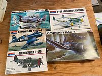 Five vintage model airplane kits in original boxes displayed on wooden surface. Boxes feature detailed illustrated artwork of WWII planes Wildcat, Corsair (two variants), Thunderbolt, and P-38 Lockheed Lightning.