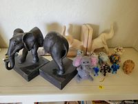 Photo showing a pair of dark metal elephant bookends with rectangular bases, placed next to cream-colored abstract elephant bookends, a plush elephant figurine, and several small elephant figurines and animal figures.