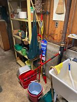 Photo showing a red mop with mop bucket, a grey upside-down bucket, several gardening tools including a garden hoe and broom, a hanging green cloth bag and various plastic bins on a shelf in a garage or workspace.