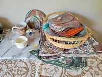 Overview of lot showing wicker basket on top of placemats, mugs, flower arrangement, and decorative plate on gray marble surface.