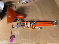 Full view of folded orange desk lamp showing the lamp shade, articulated arm with springs, and attached power cord with plug.
