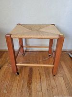 Front view of the vintage wooden stool showing woven reed seat and caster-equipped legs.