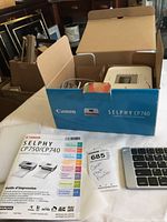 Open box showing Canon Selphy CP740 compact photo printer with original blue box and user guide manual.