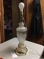 Full view of the clear pressed glass lamp base with brass accents, beige cord, and no lampshade, standing on a white surface.