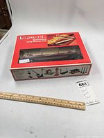 Photo of the Lionel Operating Log Dump Car 6-9335 in the original box featuring the car visible through the plastic display window on top of a white surface with a ruler nearby for scale.