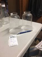 Pair of clear glass hurricane lamp shades on white table with a pen for scale. Two matching transparent glass shades, approximately 6.5 inches tall with 2 inch bases.