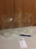 Three clear glass double end hurricane lamp shades arranged side by side with a pen for scale, showing size and shape.