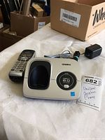 Photo showing Uniden cordless phone base unit, cordless handset, power adapter, and MaxSold tag labelled 'Uniden Cordless Phone'. Tightly arranged on a table with no other items included.