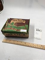 Box of toy plastic log cabin with rustic fence and tree, labeled and measured with ruler