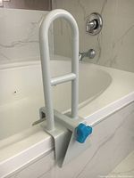 White metal bathtub side aider clamped on tub edge, showing blue plastic tightening knob and vertical support handle with horizontal grip bar.
