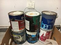 Six cans of paints in a cardboard box: three full gallon cans on the back row and three half gallon cans in front; brands include Dulux, Ben, Behr, and Evolution.