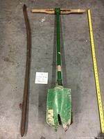 Green hand auger with wooden cross-handle on a concrete floor next to a rusty curved steel rod and a yellow measuring tape.