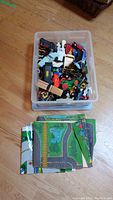 Plastic bin holding assorted toy cars and parts with two folded street-themed play mats placed beside it on floor