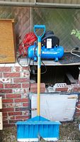 Mastercraft blue air compressor with red coiled hose and plastic snow shovel with wooden handle standing upright against brick background.
