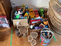 Boxes containing various toys, wooden blocks, toy trucks, and musical toys beside wicker doll chairs and bucket.