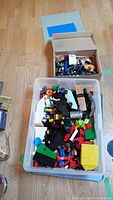 Top view of large plastic bin filled with assorted Lego blocks and a cardboard box with additional Lego pieces.