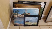Nine pieces of framed and unframed original art stacked and arranged on floor, including lighthouse painting and smaller framed coastal scene.