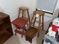 Four stools grouped together: two taller wooden round stools, one small red folding stool, one short dark wooden step stool.