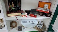 Shelf display showing an Asian miniature folding lacquer screen, two ceramic figurines, a dark green pottery bowl, a rectangular ceramic picture frame, a red tea box, and a set of sake cups.