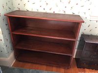 Front and angled views of a reddish-brown solid wood bookcase with three shelves, showing scratches and wear.