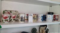 Shelf showing a collection of mugs and teacups with diverse designs including floral, bird, cows, nautical motifs and a floral plastic tray.