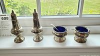 Two sterling silver salt and pepper shakers and two salt cellars with blue glass inserts arranged on a windowsill showing the full set.