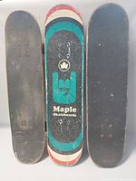 Three vintage skateboards shown standing vertically. Middle and left with plain black grip tape, the right one has black grip tape with a colorful Maple Skateboards hand sign graphic underneath.