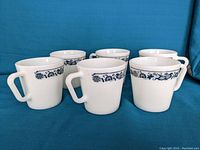 Six white Pyrex diner mugs with blue floral pattern around rim displayed on blue surface, side view showing handles.