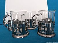 Eight clear glass cups with shiny metal holders arranged in a semi-circle showing the metal cutout details and handles.