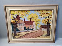 Full view of signed framed painting depicting rural scene with houses, trees with yellow leaves, and dirt road.
