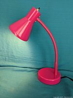 Pink adjustable gooseneck desk lamp shown from a slight side angle against a blue fabric background.