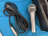Silver handheld dynamic microphone with black on/off switch and long attached cable with XLR connector, placed next to open padded case on blue background.