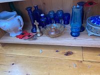 Shelf with various blue glass bottles, a white floral porcelain jug, small gold-toned bell, iridescent bowl, and other small glassware.