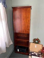 Front view of wooden storage cabinet showing two front doors with open middle and lower shelves. Cabinet positioned against wall near window with curtain.