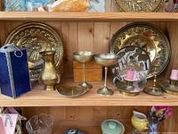 Shelf with multiple brass decor items including plates, goblets, vase, trays, and candle holders with pink candles. Also includes a blue glass lantern on the side.